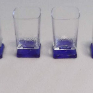 May include: Four clear glass shot glasses with blue square bases. The glasses are square shaped and have a textured surface.