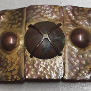 May include: A vintage, hammered metal belt buckle with a circular, copper-toned center piece. The center piece has a cross-shaped design and is surrounded by a raised, textured border.