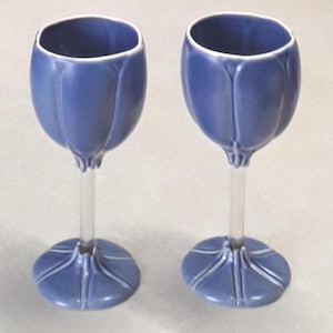 May include: Two blue tulip-shaped wine glasses with clear stems and round bases. The glasses have a smooth, glossy finish and a white rim. The bases have a decorative design. The glasses are likely for serving wine or other beverages.
