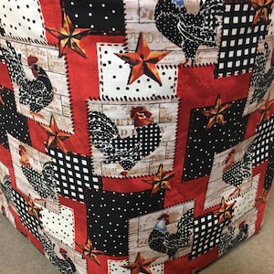 May include: A red, black, and white fabric cover with a patchwork design featuring roosters, stars, and a woodgrain pattern. The cover is made of cotton and has a quilted texture.