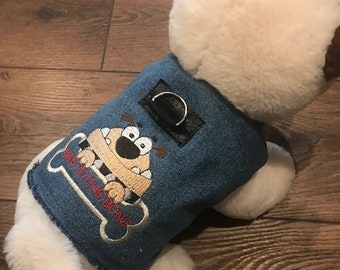 May include: A white stuffed dog wearing a blue denim vest with an embroidered cartoon dog and the text "Wag to the Bone".