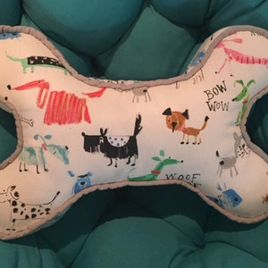 May include: White dog bone shaped pillow with a gray border. The pillow is covered in a white fabric with a pattern of colorful cartoon dogs and the words "Bow Wow" and "Woof".