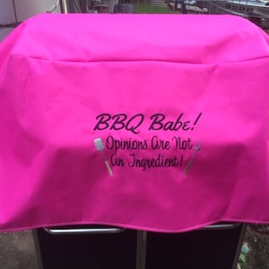 May include: Pink grill cover with the text "BBQ Babe! Opinions are not an ingredient!" embroidered on it.