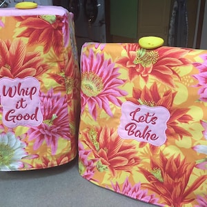 May include: Two yellow and pink floral patterned kitchen appliance covers with embroidered text. The left cover says "Whip it Good" and the right cover says "Let's Bake".