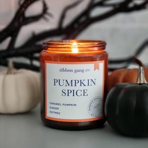 May include: A lit candle in an amber glass jar with a white label featuring the text ribbon gang co. PUMPKIN SPICE, CARAMEL PUMPKIN, GINGER, NUTMEG, and a circular logo. The label has an orange border and a small orange ribbon icon at the top.