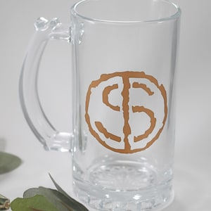 May include: A clear glass beer mug with a handle. The mug has a gold-colored circle with the letters "CS" inside.