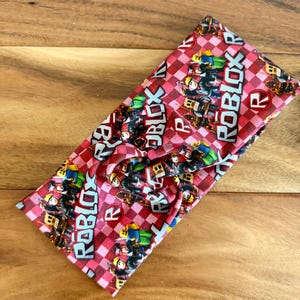 Peut inclure: Un bandeau rouge et blanc à carreaux avec le mot "ROBLOX" en lettres blanches. Le bandeau présente un motif répété de personnages et de logos Roblox. Le tissu est noué en haut.