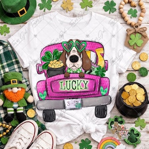May include: White t-shirt featuring a pink truck with a cartoon dog wearing heart-shaped green glasses. The truck bed is filled with a pot of gold, shamrocks, and the word "LUCKY". The date "3-17" is on the license plate.