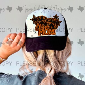 Puede incluir: Gorra de camionero negra y blanca con una malla blanca en la parte posterior. La parte delantera de la gorra presenta un pompón de animadora naranja y negro con el texto "Cheer Mom" en naranja.