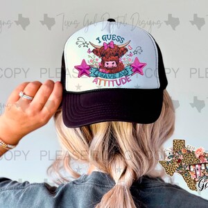 May include: A white and black trucker hat with a pink and white cow graphic and the text "I guess I have an attitude". The hat is being worn by a person with blonde hair. A separate sticker of a leopard print Texas outline with the text "Texas Gal" is visible in the background.