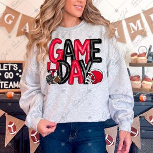 Puede incluir: Una sudadera gris claro con la frase "GAME DAY" en rojo y negro. El diseño incluye un balón de fútbol americano, un casco y pompones. Esta sudadera forma parte de una colección de ropa para el día del partido.