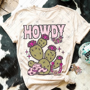 May include: A beige t-shirt with the word "HOWDY" in pink letters and a cactus with pink flowers, a butterfly, a horseshoe, and a pink cowboy hat.