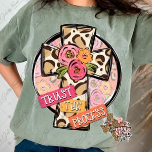 Puede incluir: Camiseta verde salvia con un diseño de cruz con estampado de leopardo. La cruz está adornada con flores rosas y naranjas y las palabras "TRUST THE PROCESS". También se ve un gráfico en forma de Texas con detalles florales y de estampado de leopardo.