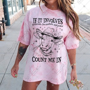 May include: A pink tie-dye t-shirt with a black and white illustration of a cow wearing a hat and the text "If it involves backroads, flea markets, and junkin' Count me in".