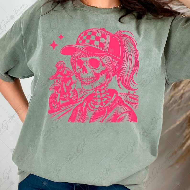 May include: A green t-shirt with a pink graphic of a skeleton wearing a checkered cap and riding a dirt bike. The skeleton has a ponytail and is wearing a racing jacket. The graphic is in a vintage style.