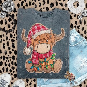 May include: A grey crew neck t-shirt with a cartoon Highland cow wearing a Santa hat and a red plaid scarf. The cow is decorated with a string of colourful Christmas lights. The design is printed on a soft, comfortable fabric.