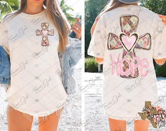 May include: Cream-colored t-shirt featuring a cross design with a pink heart in the center. The cross is decorated with floral and patterned elements. The word "HOPE" is printed in pink on the back of the shirt. The shirt has short sleeves.