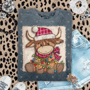 May include: A grey t-shirt with a cartoon Highland cow wearing a red plaid Santa hat and scarf, and a string of colourful Christmas lights. The cow is on a burlap-textured square.