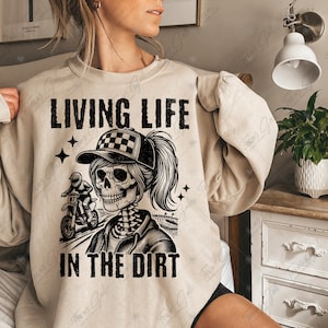 May include: A beige sweatshirt with a black graphic of a skeleton woman riding a dirt bike. The text "LIVING LIFE IN THE DIRT" is printed on the sweatshirt.
