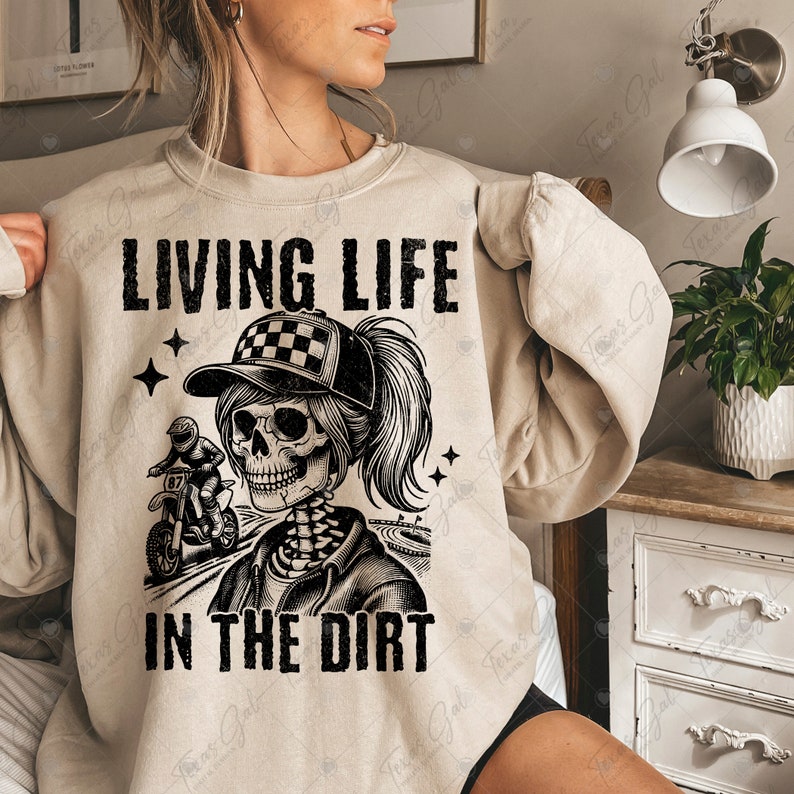 May include: A beige sweatshirt with a black graphic of a skeleton woman riding a dirt bike. The text "LIVING LIFE IN THE DIRT" is printed on the sweatshirt.