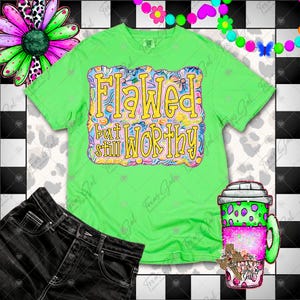 Op de afbeelding: Een limoengroen t-shirt met de woorden "Flawed but still WORTHY" in een kleurrijk bloemmotief. De afbeelding bevat ook een zwarte spijkerbroek en een koffiekopje met een roze en groen ontwerp.