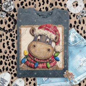 May include: A gray t-shirt featuring a whimsical illustration of a cow wearing a Santa hat and a red plaid scarf adorned with Christmas lights. The cow's face is textured, giving it a rustic appearance. The design is set against a burlap-like background.