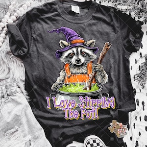 May include: Dark gray t-shirt featuring a raccoon wearing a purple witch hat and orange overalls, stirring a green potion in a cauldron. The text "I Love Stirring The Pot!" is below the image.