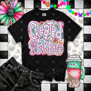 May include: Black t-shirt with the text "Created for a Purpose" in a colorful design. The image also includes a pair of black jeans and a coffee cup with a Texas-themed design. The background has a black and white checkered pattern.