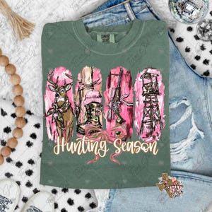 May include: Green Comfort Colors t-shirt with a pink and brown hunting-themed graphic. The graphic includes a deer, boots, a rifle, and a hunting stand, with the words "Hunting Season" in a cursive font.