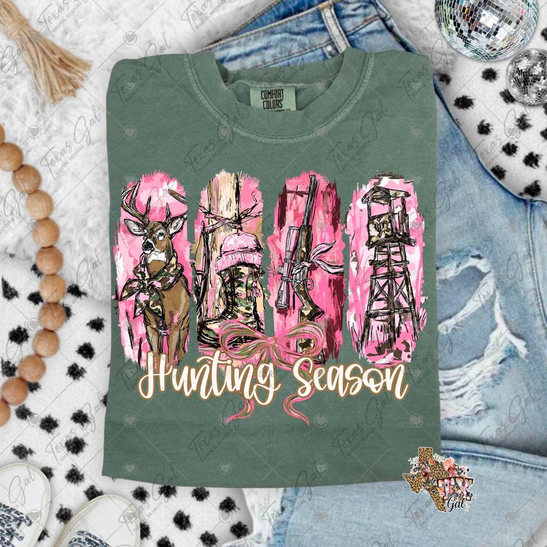 May include: Green Comfort Colors t-shirt with a pink and brown hunting-themed graphic. The graphic includes a deer, boots, a rifle, and a hunting stand, with the words "Hunting Season" in a cursive font.