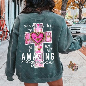May include: A dark green sweatshirt with a pink and white cross design. The cross has a heart in the center and the text "saved by his amazing grace" is printed below it.