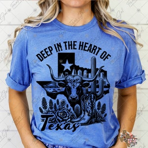 Op de afbeelding: Lichtblauw t-shirt met de tekst "DEEP IN THE HEART OF Texas". De afbeelding toont een longhorn, de omtrek van Texas, een cactus en cowboylaarzen in zwart.
