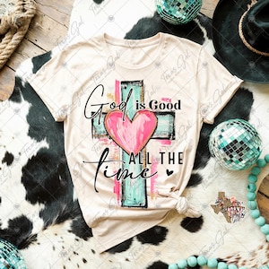 May include: A beige t-shirt with a graphic design of a cross with a heart in the center. The text "God is Good All The Time" is written on the shirt.