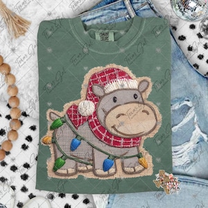 以下が含まれることがあります： セージグリーンのスウェットシャツ。赤いチェック柄の帽子とスカーフを身につけ、クリスマスライトで飾られた刺繍の入ったカバが特徴です。カバはジュート風のパッチの上にいます。スウェットシャツには「COMFORT COLORS」の文字が入っています。