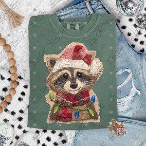 May include: Sage green Comfort Colors sweatshirt featuring an embroidered raccoon wearing a Santa hat, a red plaid scarf, and a string of Christmas lights. The raccoon has brown and tan fur with black eyes and a smiling expression. The sweatshirt has the text "Texas Gal" printed on it.