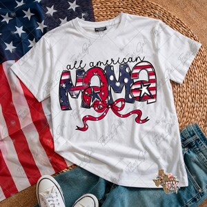 May include: White t-shirt with the text "all american MAMA" in red, white, and blue, with a red bow. The shirt is paired with blue jeans and white sneakers, with an American flag in the background.
