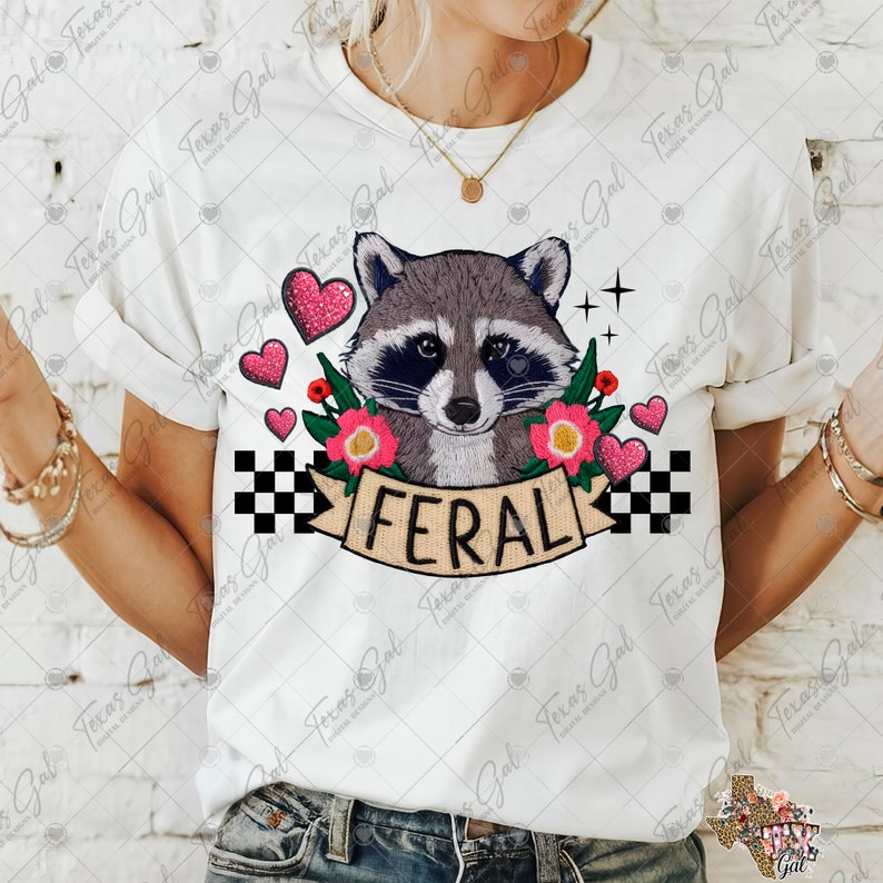 May include: White t-shirt with a graphic of a raccoon with pink flowers and the word "FERAL" in black lettering. The raccoon is embroidered with a gray and black color scheme.
