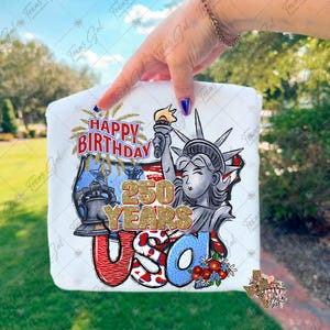 May include: White t-shirt featuring a graphic design celebrating the USA's 250th anniversary. The design includes the Statue of Liberty, fireworks, and the text "HAPPY BIRTHDAY 250 YEARS USA" in red, blue, and gold glitter.
