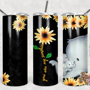 May include: A black and yellow stainless steel tumbler with a sunflower design and the text "you are my sunshine". The tumbler is shown with a straw and a gray elephant with a baby elephant.