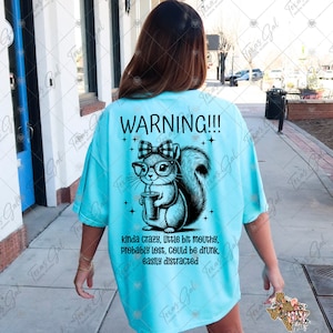 May include: A turquoise blue t-shirt with a black and white illustration of a squirrel wearing glasses and a bow. The text "WARNING!!!" is above the squirrel and the text "kinda crazy, little bit mouthy, probably lost, could be drunk, easily distracted" is below the squirrel.