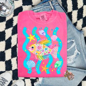 May include: A pink t-shirt featuring a colorful fish design with floral patterns, seashells, and starfish. The fish is surrounded by blue wavy lines, creating an underwater theme. The shirt is made of a soft material.
