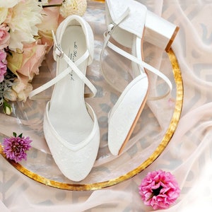 K&ouml;nnte beinhalten: Ein Paar wei&szlig;e Satin-Brautschuhe mit zarten Spitzendetails und einem kleinen Blockabsatz. Die Schuhe befinden sich auf einem goldenen Teller mit einem Blumenstrau&szlig; im Hintergrund.