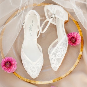 May include: A pair of white, floral lace bridal flats with ankle straps and silver buckles. The shoes are displayed on a gold-rimmed plate with pink flowers and a sheer white veil.