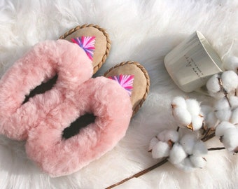 May include: Pair of pink fluffy sheepskin slippers with brown leather trim and colourful embroidery. The slippers are on a white fluffy surface.