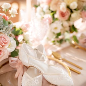 May include: A pair of white, lace-covered wedding flats with ankle straps, resting on a gold-rimmed plate with a pink napkin. The table is decorated with a bouquet of pink and white flowers and gold cutlery.