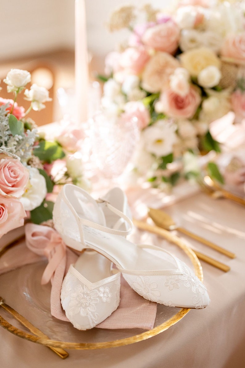 May include: A pair of white, lace-covered wedding flats with ankle straps, resting on a gold-rimmed plate with a pink napkin. The table is decorated with a bouquet of pink and white flowers and gold cutlery.