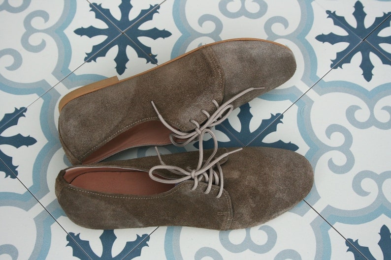 May include: A pair of brown suede lace-up shoes with a rounded toe. The shoes are on a white and blue patterned tile floor.