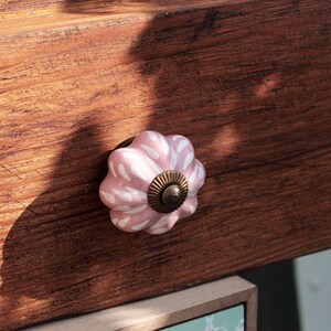 Puede incluir: Un pomo de caj&oacute;n de cer&aacute;mica rosa con un patr&oacute;n floral blanco y un centro de lat&oacute;n. El pomo est&aacute; montado en un frente de caj&oacute;n de madera.