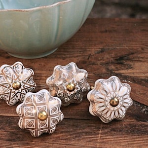 Könnte beinhalten: Vier Schubladenknöpfe aus Keramik mit einer gewellten, blumenartigen Form. Jeder Knopf weist ein einzigartiges weißes und goldfarbenes geometrisches oder florales Muster sowie einen zentralen goldfarbenen Metallschraubenkopf auf.