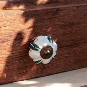 May include: White ceramic drawer knob with a green leaf design. The knob is mounted on a wooden drawer front.